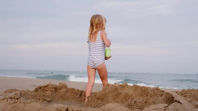 Wet three years old girl playing with water and sand on beach at seaside. Vacations with kid, tourism concept.