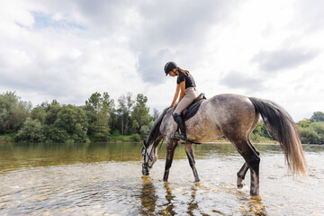 Girl riding gray horse down the calm river water with forest greenery reflections from the...