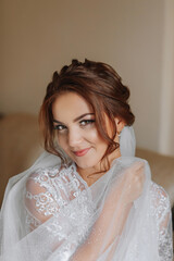 Fototapeta na wymiar The morning of the bride in a white robe and an elegant hairstyle. An adult girl sits on a chair and fastens an earring, looking into the camera lens and smiling