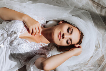 A beautiful young bride in a white robe poses for a photographer, lying on the floor, on a veil. Wedding photography, close-up portrait, chic hairstyle.