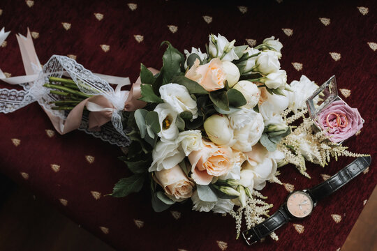 Photo of wedding accessories. Luxurious bouquet of the groom. Gold rings on a stand with flowers, a black classic watch on a burgundy background.
