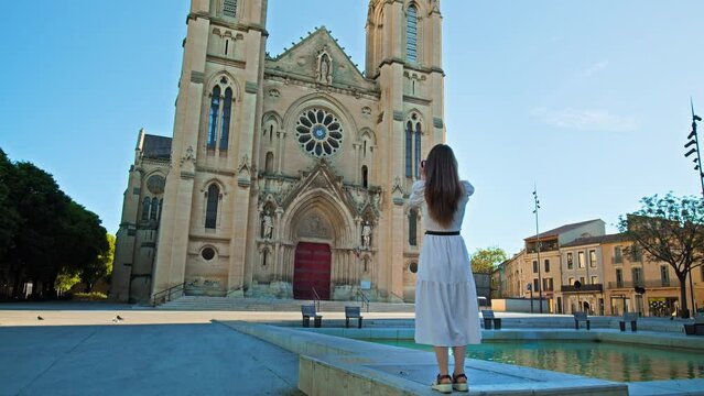 A girl tourist visits catholic church in the neo-Gothic style in Nimes, France. Young woman taking pictures of an old historical building with two bell towers on the facade surmounted by high Spiers.