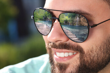 Man in sunglasses on sunny day outdoors. Lake with wooden bridge reflecting in lenses