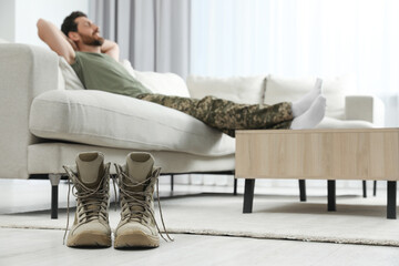 Soldier relaxing on sofa in living room, focus on pair of combat boots. Military service