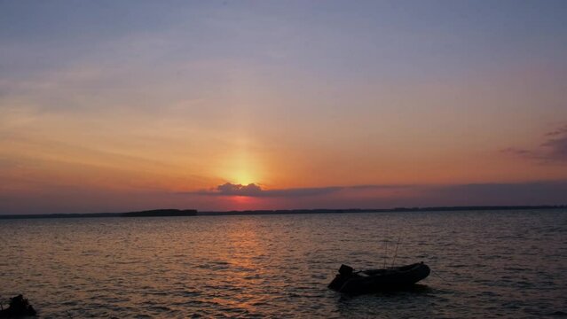 Anchored boat silhouette. Inflatable motorized boat with fishing rods swaying on tranquil river waves at sunset.