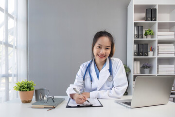 Asian doctor young beautiful woman smiling using working with a laptop computer and her writing something on paperwork or clipboard white paper at hospital desk office, Healthcare medical concept