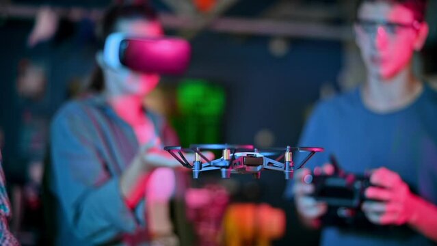 Teens doing experiments in robotics in a laboratory. Boy with controller and girl in VR headset controlling a flying drone using her hand. Red and blue illumination. Slow motion