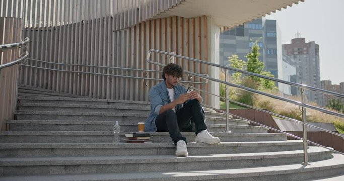 Young indian guy student holding mobile phone and typing message while sitting on a stairs outdoor