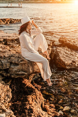 woman sea sunset. woman in a white pantsuit and hat is sitiing on the beach enjoying the sea. Happy summer holidays