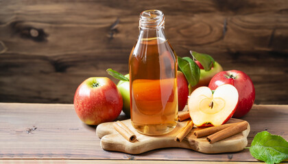 Apple cider in glass bottle with cinnamon sticks and fresh apples on cutting board, on wooden...