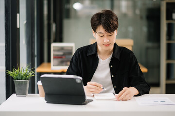 Young attractive Asian man smiling thinking planning writing in notebook, tablet and laptop working from home, looking at camera at office .