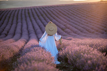 Happy woman in a white dress and straw hat strolling through a lavender field at sunrise, taking in the tranquil atmosphere.