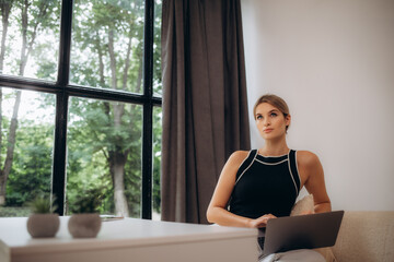 Happy beautiful woman watching videos or enjoying entertainment content on a laptop sitting near windows in the house.