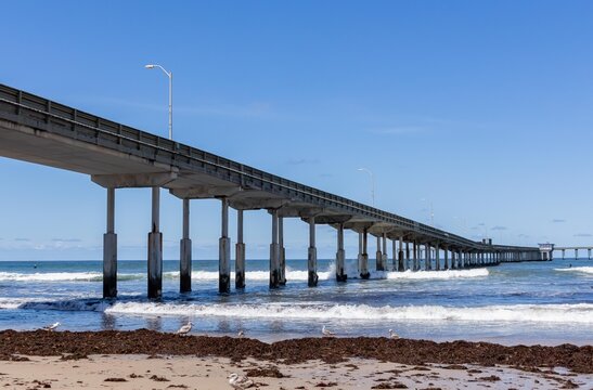 Concrete pier crossing from left to right in distance with crashing waves and surfers, horizontal