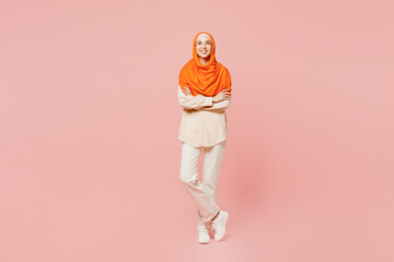 Fototapeta premium Full body smiling happy young arabian asian muslim woman wears orange abaya hijab hold hands crossed folded look camera isolated on plain pink background. Uae middle eastern islam religious concept.