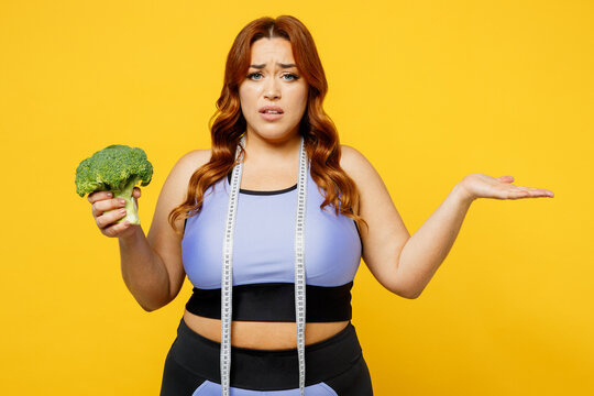 Young confused overweight plus size big fat fit woman wears blue top warm up train hold tape measure eat broccoli spread hand isolated on plain yellow background studio home gym Workout sport concept