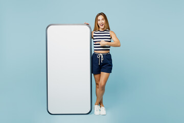 Full body young woman she wears striped tank shirt casual clothes point on big huge blank screen mobile cell phone smartphone with workspace area isolated on plain pastel light blue cyan background.