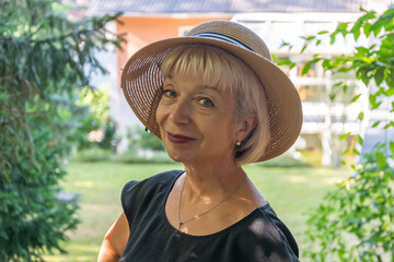 Street portrait of an elderly woman wearing a straw hat, 60-65 years old, against the background of a European country house and a verdant garden.