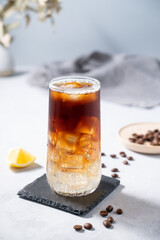 Coffee with tonic in a tall glass with ice and lemon on a light blue background with coffee beans and fruit. Trendy summer craft refreshing drink.