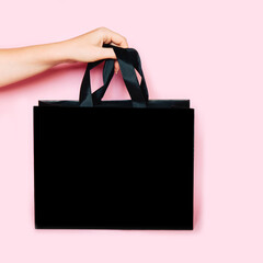Woman holding shopping black paper bag with silk handle mockup, on pink background. Empty gift pack...