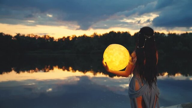 Fantasy woman holding moon magic light glowing planet in hands hugs back rear view. Girl night elf beauty face, river nymph blue dress, water dark sky clouds lake. Lady magician fairy queen art. 4k