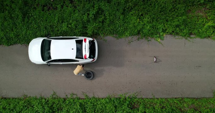 Aerial view, a young man changes a wheel on a car on the side of the road. Spare wheel. An emergency sign. Top view. Emergency stop