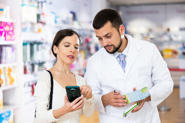 Young adult woman customer showing man pharmacist electronic prescription in her smartphone and...