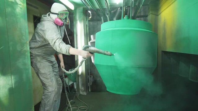 Industrial painting process in chamber. Painter spraying green paint on detail in special booth. Technician in safety wear work at industrial manufacture. Master paint parts in specialised workshop