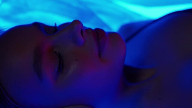 Meditation, cheerful young woman with headphones lies in the sensory deprivation chamber, blue neon light, relax in spa, portrait.