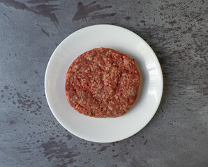 Single raw hamburger patty on a white plate top view atop a gray mottled background.