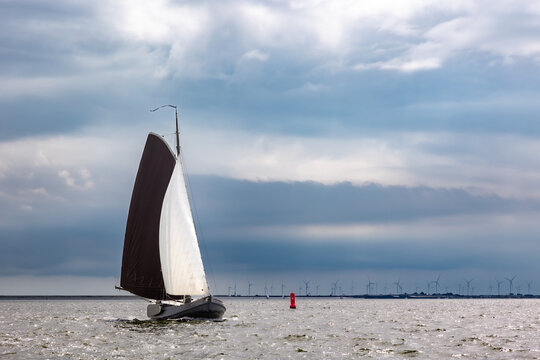 Traditional wooden Dutch sailboat with brown and white sails sailing under stormy, omnious, thunder skies along the coast on the sea. Bright red buoy and windmills on the horizon.