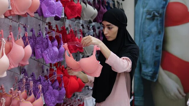 Muslim woman customer, female client in lingerie store chooses pink cute bra for everyday wear. Islamic woman in hijab in lingerie boutique chooses underwear, bras. Choosing and buying clothes in mall