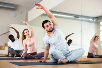 Fotobehang Young bearded man doing stretching exercises during group training in fitness center, sitting in lotus position, bending sideways with arm raised overhead © JackF