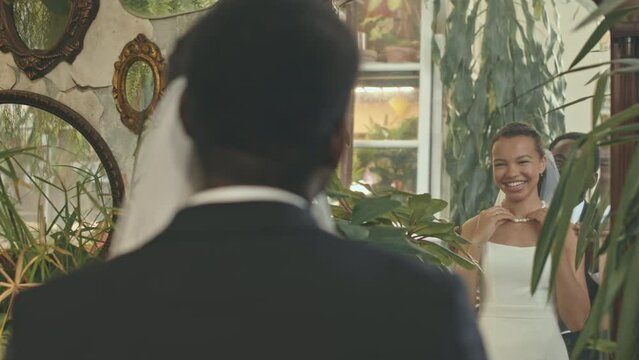 Back view of contented young Black couple in wedding costumes standing in front of full length mirror. Groom wearing beautiful pearl necklace on neck of his beloved bride