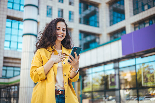 Beautiful young woman is using an app in her smartphone device to send a text message near business buildings. Young positive businesswoman with take away coffee cup in hand using mobile phone