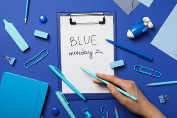Blue Monday text, stationery and hand with pencil on blue background, top view