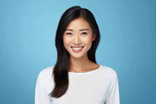 Pretty Asian Woman With Dark Hair, Wearing A White Shirt And Jumper, Smiles Pleasantly At The Camera With A Toothy Smile. Isolated Over A Blue Background.