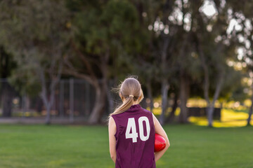 schoolgirl wearing footy jersey with football under her arm seen from behind