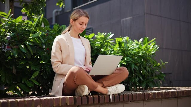 Relax freelancer in a beautiful brown suit and glasses opens laptop and starts working, typing a message sitting on a bench. IT specialist writes a piece of code, enjoying the warmth of sunny day.
