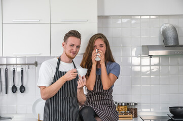 Caucasian young couple in white kitchen holding cups of coffee wearing aprons.