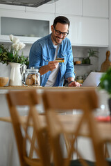 Smiling businessman paying through credit card while online shopping over laptop on kitchen counter