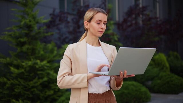 Focused freelancer in a beautiful brown suit and glasses opens laptop and starts working, typing a message sitting on a bench. IT specialist writes a piece of code, enjoying the warmth of sunny day.