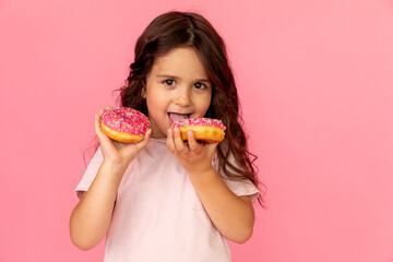 Portrait of a happy little smiling girl with curly hair and two appetizing donuts in her hands on a pink background, a place for text. Dieting concept and junk food.