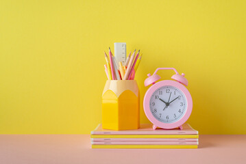 Back to school concept. Photo of school supplies on pink desktop alarm clock and pencil holder over stack of notebooks on yellow wall background