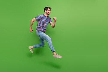 Fototapeta na wymiar Full length photo of excited positive guy wear striped t-shirt running fast jumping high empty space isolated green color background