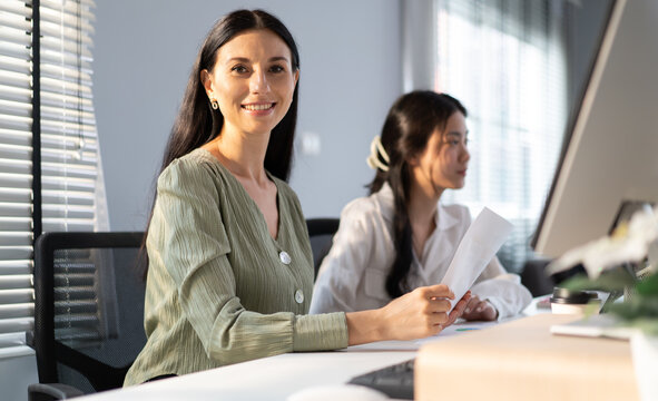 Female apprentice learning business from caucasian businesswoman in office. Professional manager teaching young Asian trainee using tablet. Portrait of confident mentor and intern talking in workplace