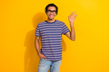 Photo of good mood pleasant man with dreadlocks dressed striped t-shirt waving palm arm in pocket isolated on yellow color background