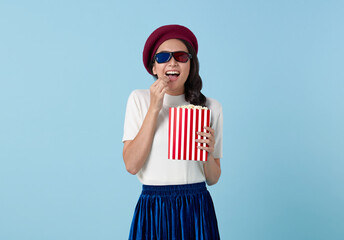 Young smiling fun cool Asian woman in 3d glasses watch movie film hold bucket of popcorn isolated on blue background studio. People cinema lifestyle concept