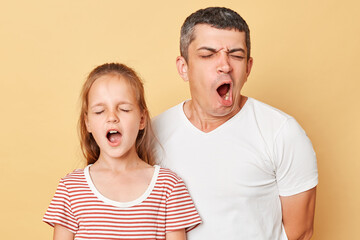 Fototapeta premium Little adorable daughter with father wearing casual striped T-shirt standing with closed eyes yawning with widely open mouth need having nap.