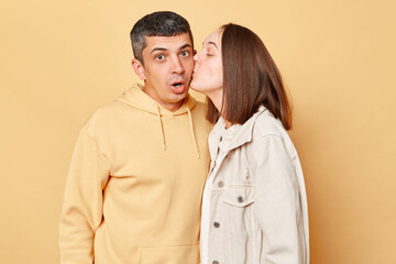 Funny young man and woman couple standing together isolated over beige background wife kissing astonished husband looking with big eyes.